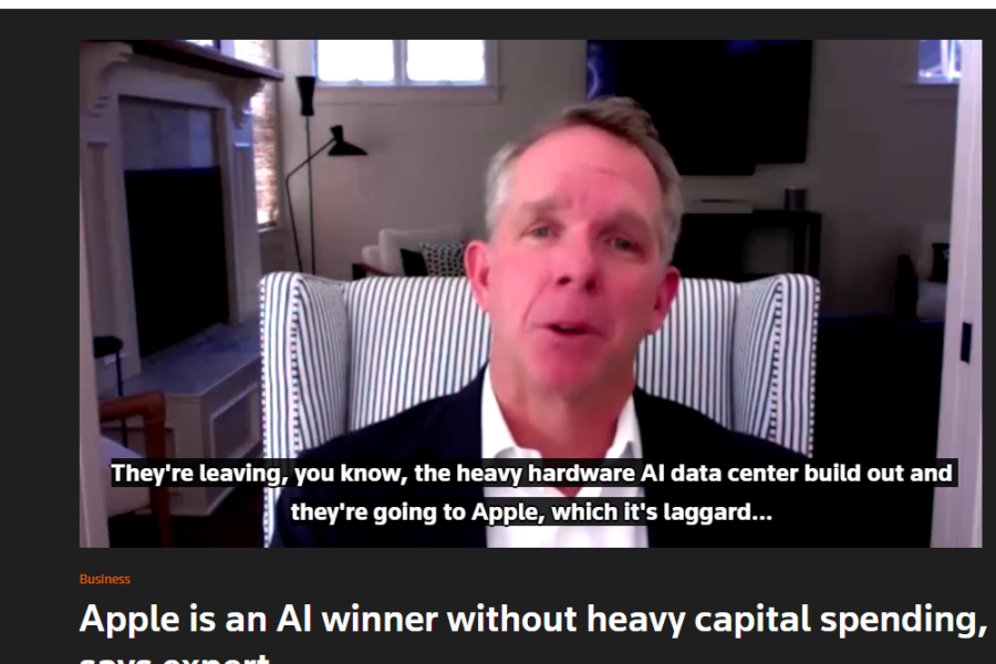 Video interview screenshot of a man in a suit speaking from a striped chair in a home office; onscreen subtitles discuss AI hardware and Apple.